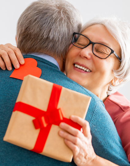 Older adult woman with glasses with arms wrapped around an older adult male, who is seen from behind. The woman is holding a red heart in her right hand and a wrapped gift in her left hand