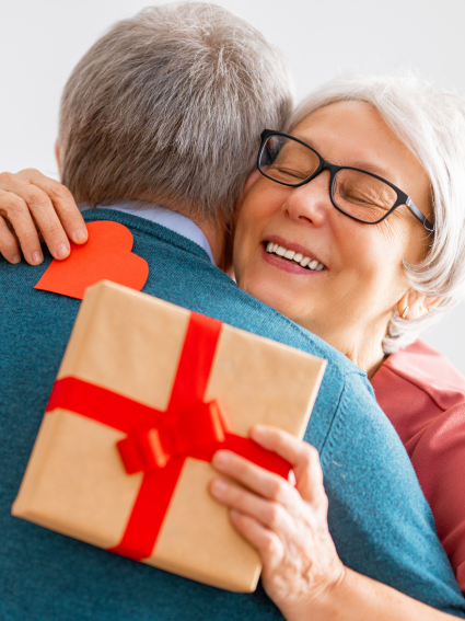 Older adult woman with glasses with arms wrapped around an older adult male, who is seen from behind. The woman is holding a red heart in her right hand and a wrapped gift in her left hand